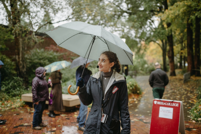 Student with a jacket standing outside with an umbrella on a rainy day at Reed