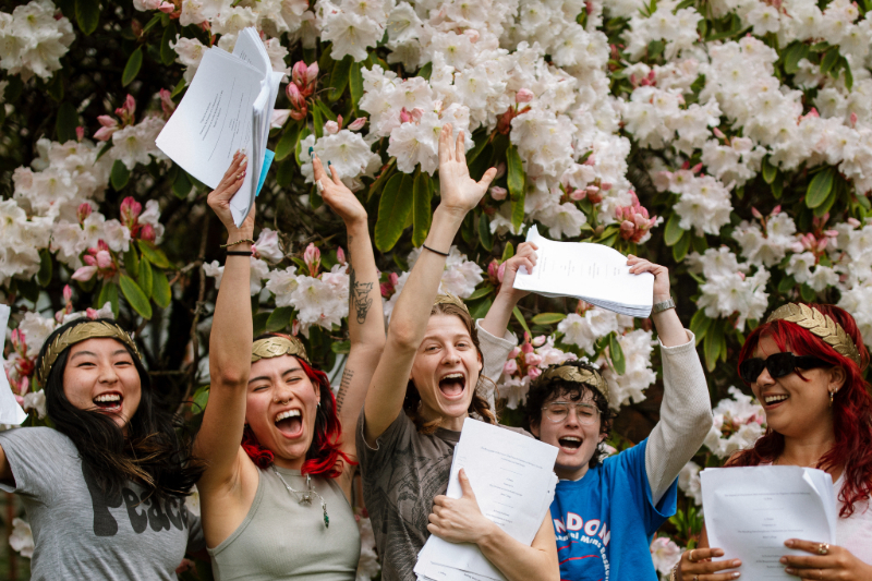 Five students wearing golden Roman laurels holding papers and celebrating